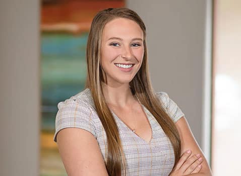 Ivy Reynolds - a young woman with long blonde hair wearing a light grey plaid dress. She is smiling directly at the camera and has her arms crossed.